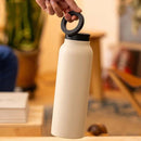 White water bottle with black lid held by a hand in a blurred indoor setting
